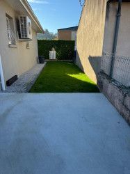 Cour arrière étroite avec terrasse en béton, pelouse verte, murs beiges et une haie au fond.