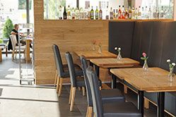 Café-Innenraum mit Holztischen, schwarzen Stühlen, Bar im Hintergrund und einer sitzenden Person in der Ferne.