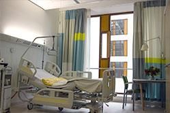 Krankenzimmer mit verstellbarem Bett, Fenster, Vorhängen und Beistelltisch.
