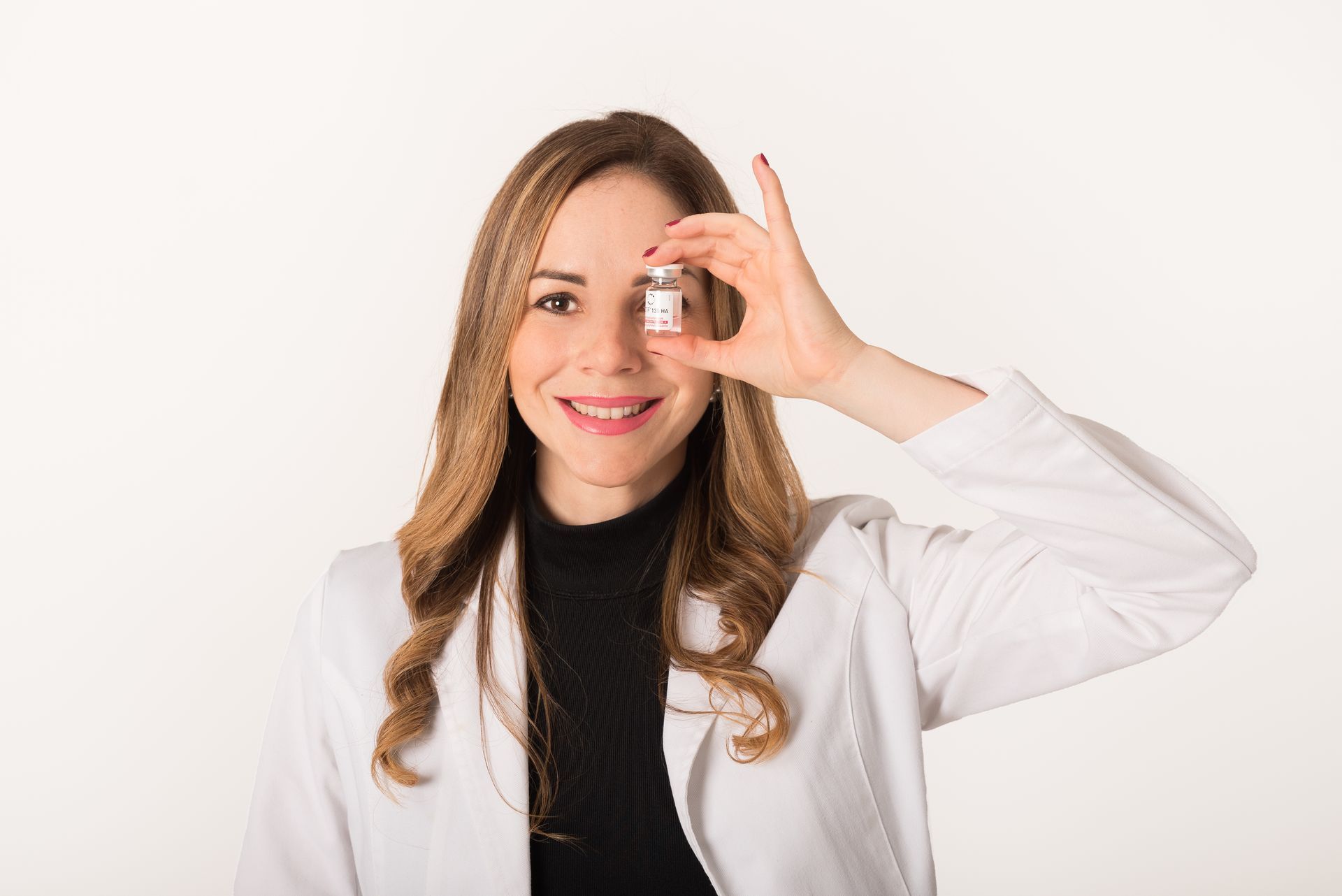Mujer con bata de laboratorio sosteniendo un frasco frente a su ojo, sonriendo.