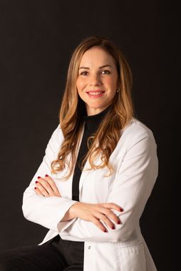 Mujer con bata de laboratorio blanca sonríe con los brazos cruzados, fondo negro.