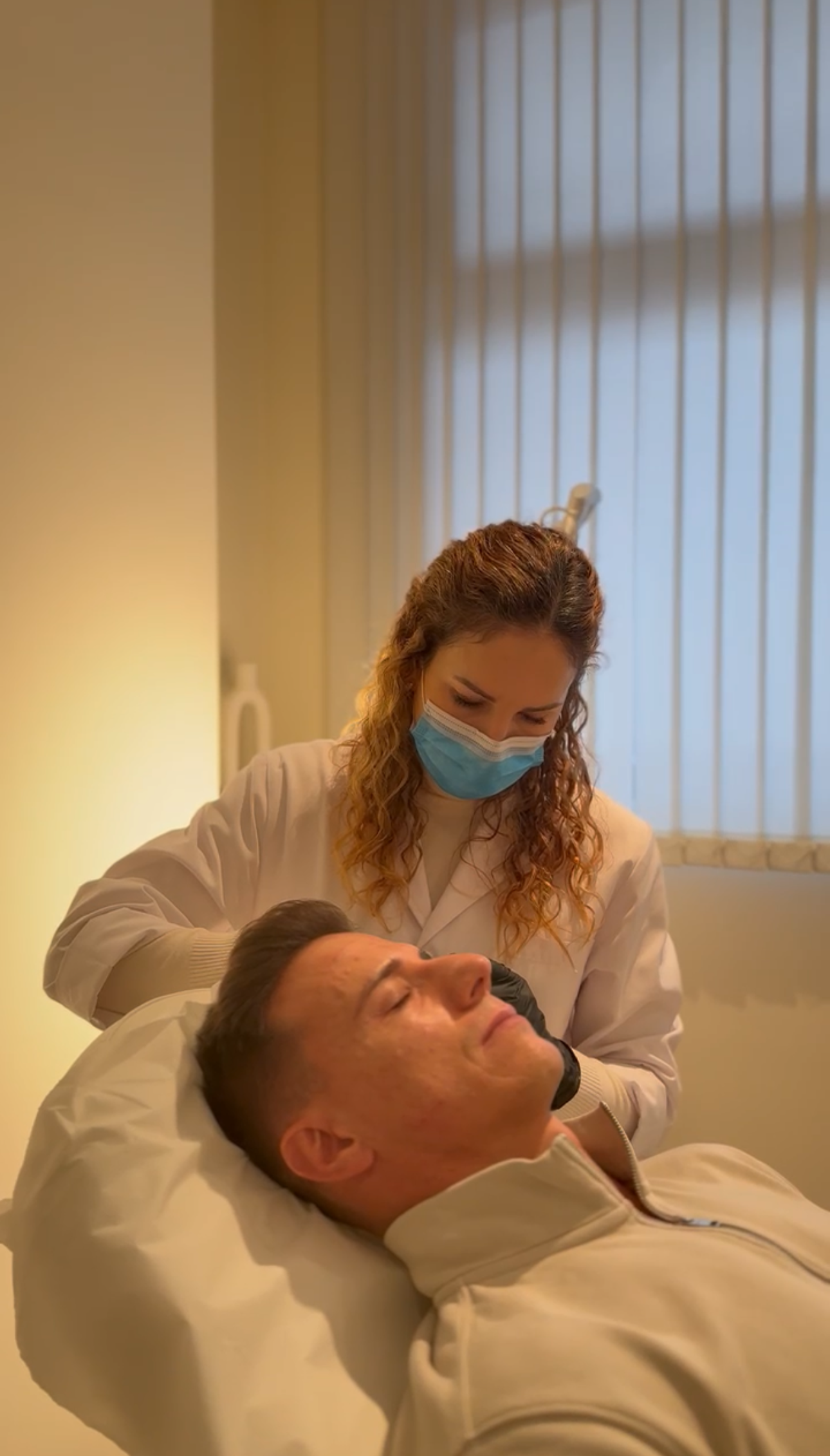 Mujer con bata de laboratorio y mascarilla realizando un procedimiento a un hombre acostado en una cama médica.