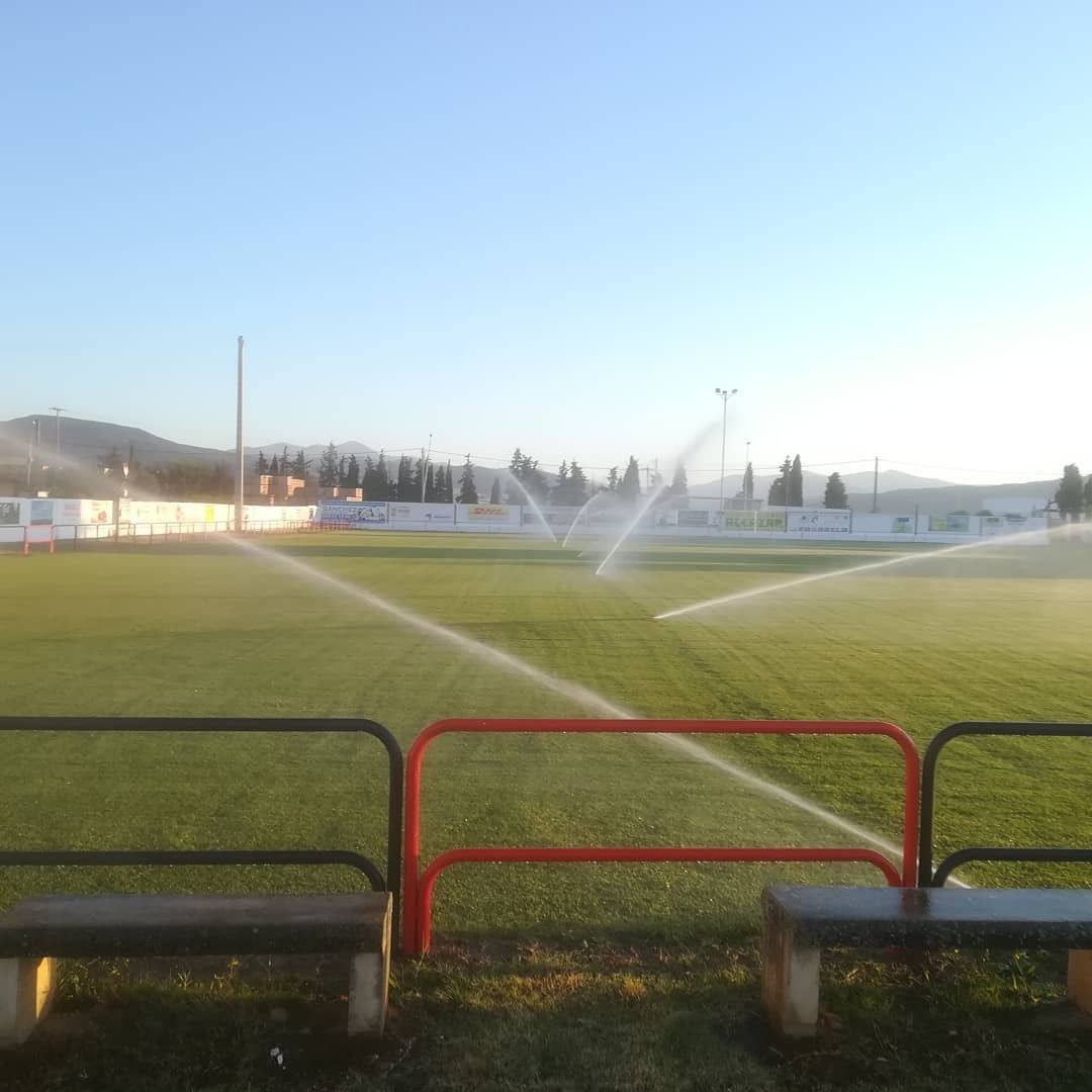 Campo de fútbol regado con aspersores; barreras rojas y negras en primer plano, montañas en la distancia.