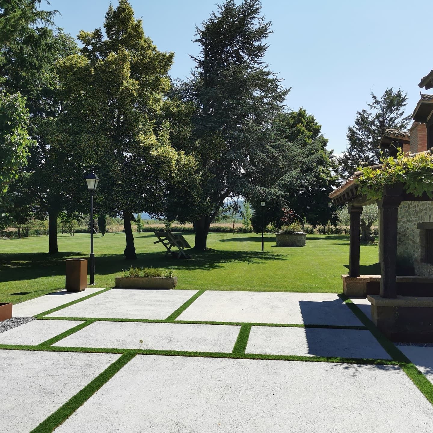 Una escena al aire libre soleada con un patio con insertos de césped, con vista a un exuberante césped verde y árboles.