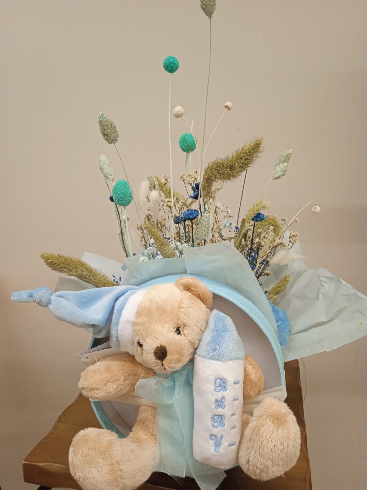 Un osito de peluche sostiene un biberón delante de un ramo de flores.