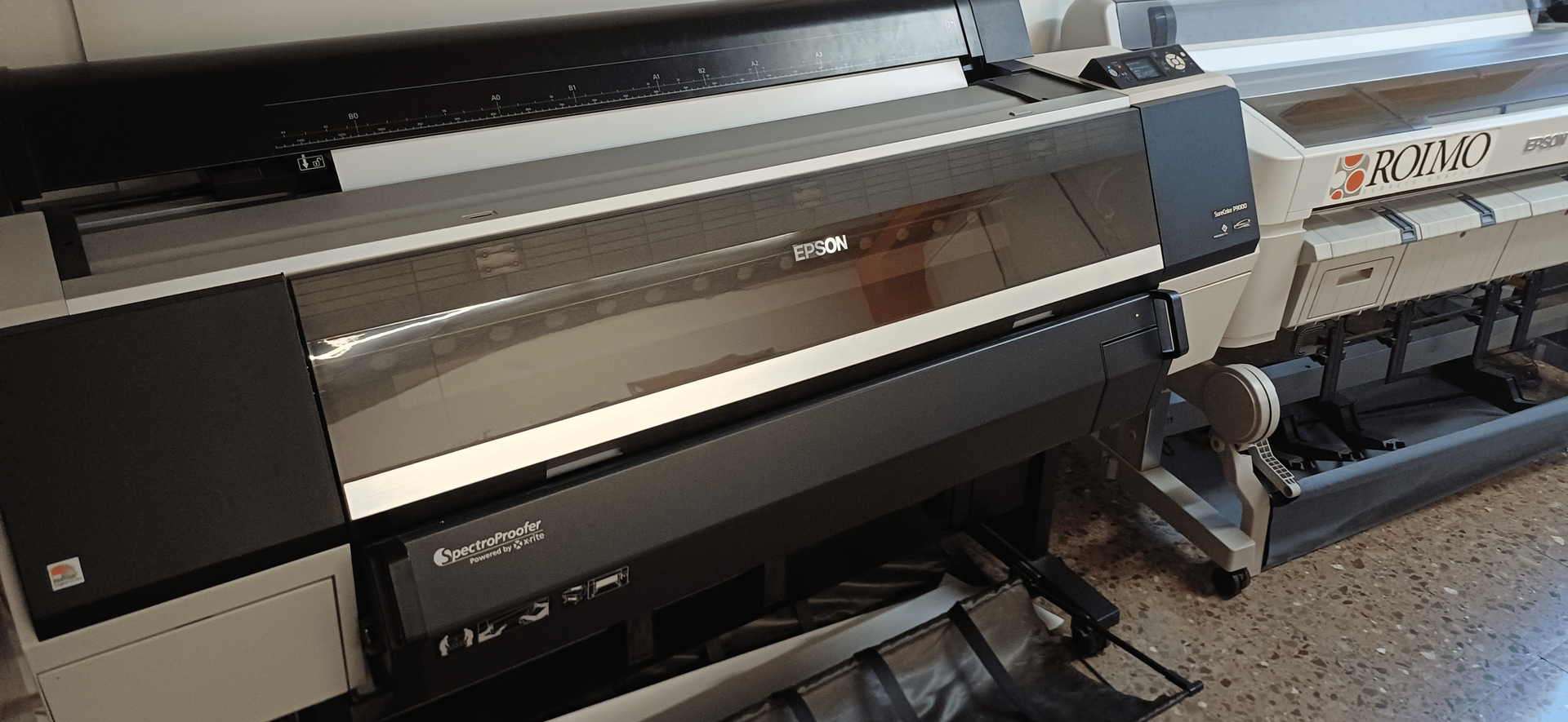 Una impresora de gran formato en una habitación bien iluminada. La impresora es negra y gris, con marcas de marca visibles.