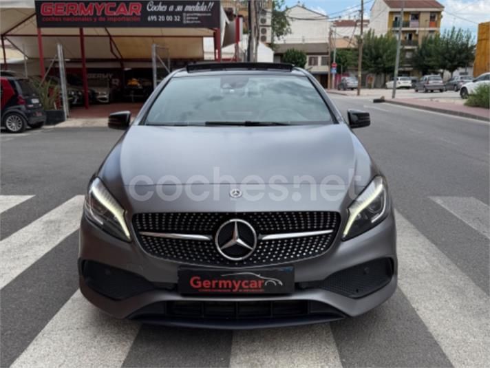 Un automóvil Mercedes-Benz Clase A de color gris estacionado en una calle con un concesionario al fondo.