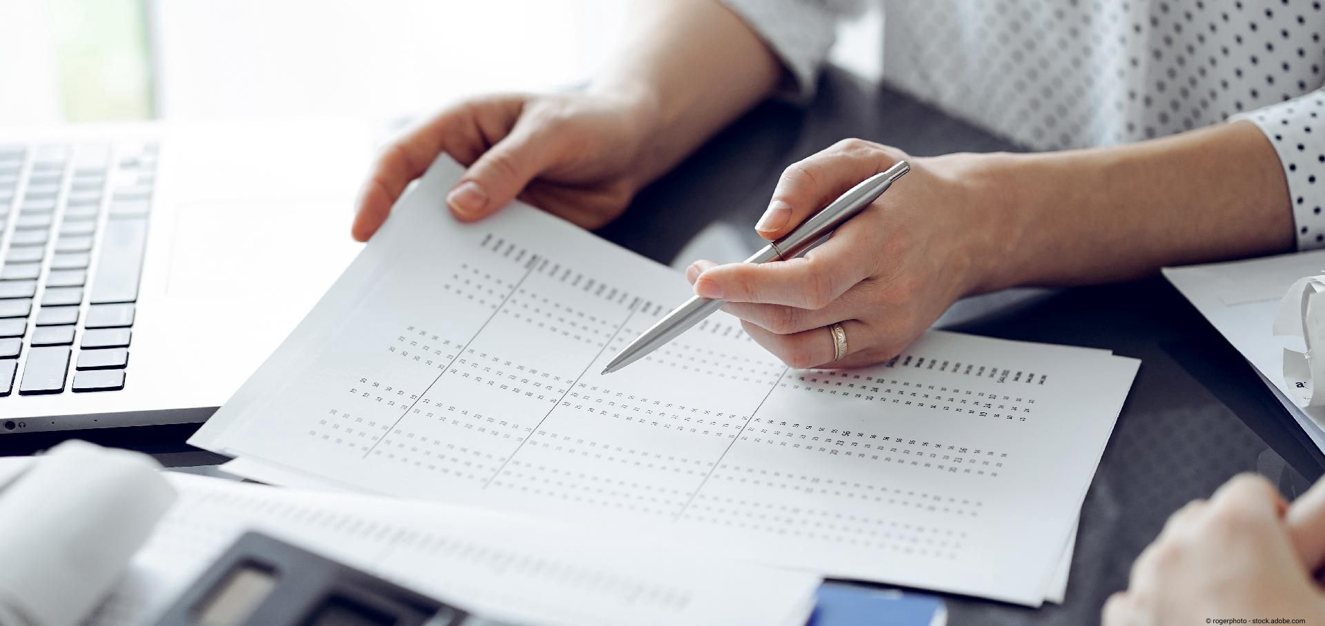 Hände, die ein Dokument mit Zahlen prüfen und einen Stift halten.