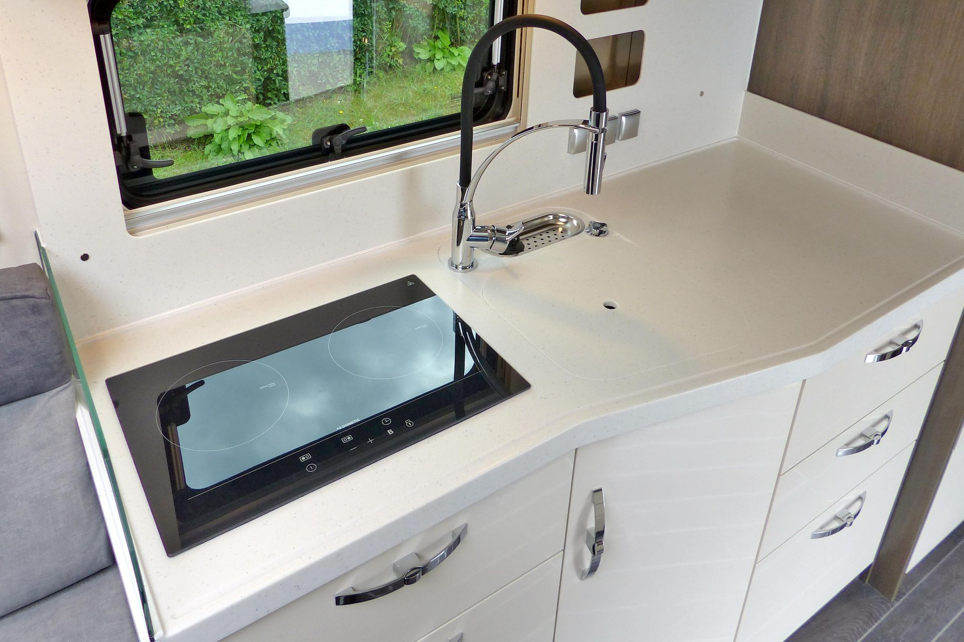 Weiße Arbeitsplatte in einer Wohnmobilküche mit Spüle, Wasserhahn, Herdplatte und Schubladen in der Nähe eines Fensters.