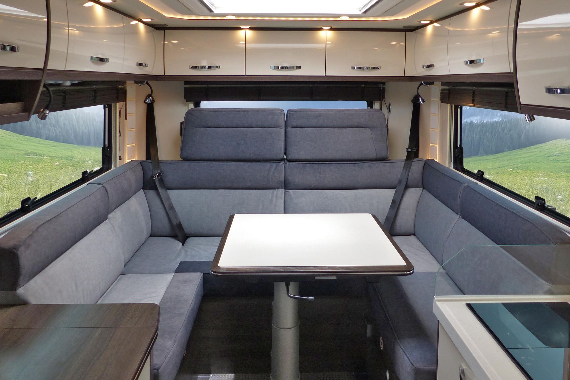 Innenraum eines Wohnmobils: graue Sitzgelegenheiten, weißer Tisch, Hängeschränke und Fenster mit grüner Landschaft.