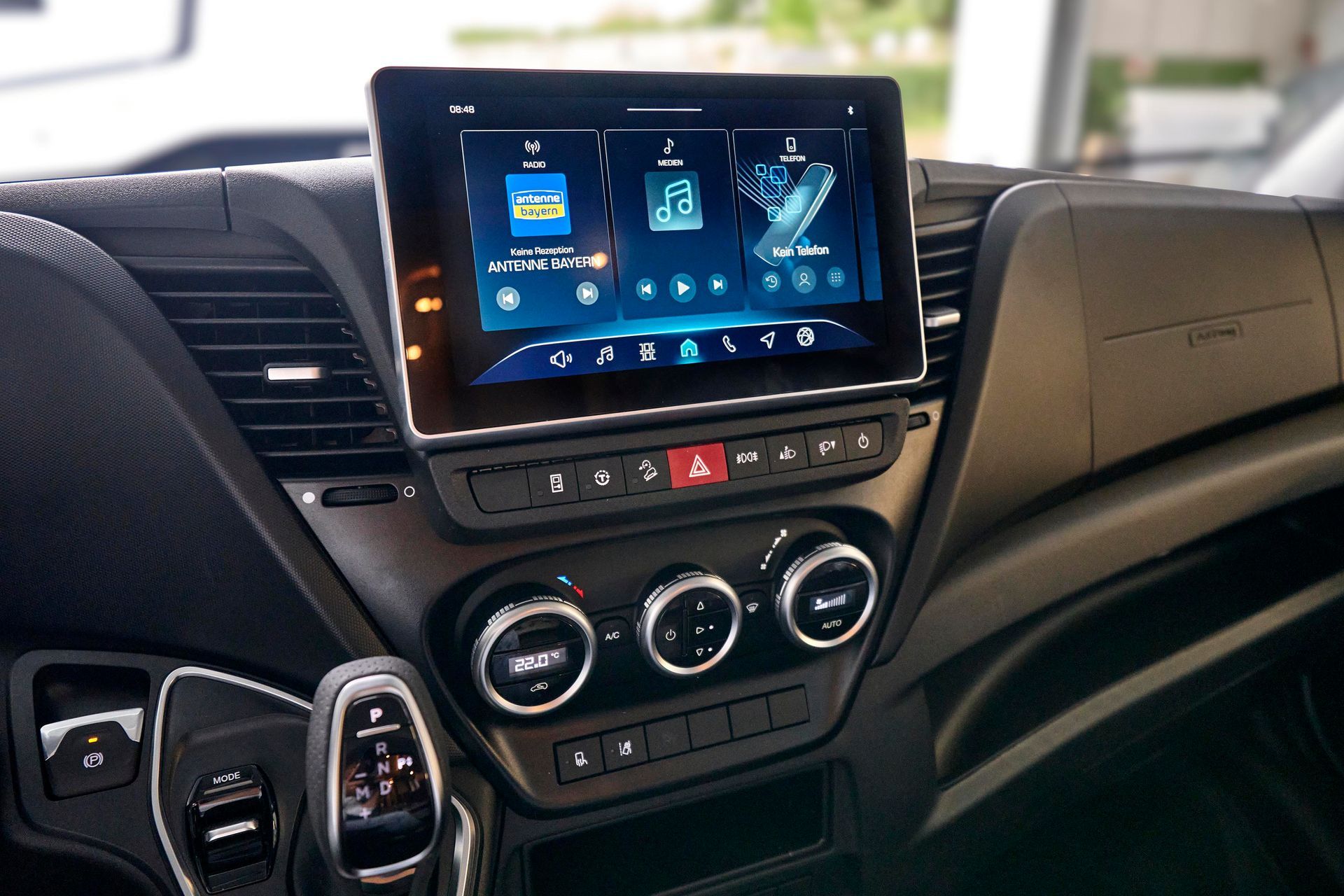 Armaturenbrett eines Fahrzeugs mit großem Touchscreen-Display. Enthält Bedienelemente für Klimaanlage, Warnblinkanlage und Gangschaltung.