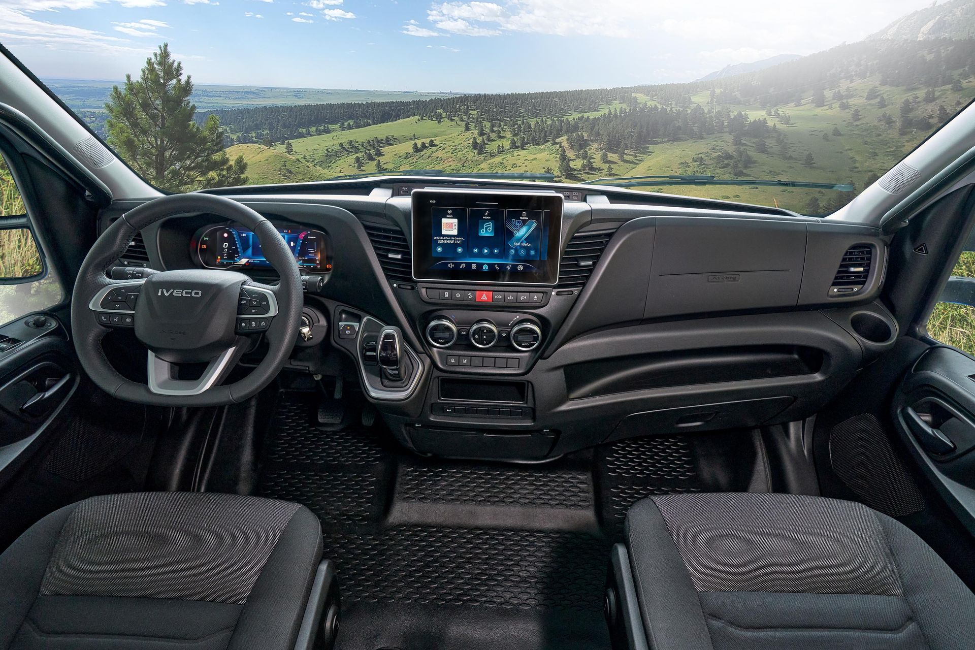 Innenraum eines schwarzen Lieferwagens mit Armaturenbrett, Lenkrad und Infotainmentsystem und Blick auf eine malerische Landschaft.