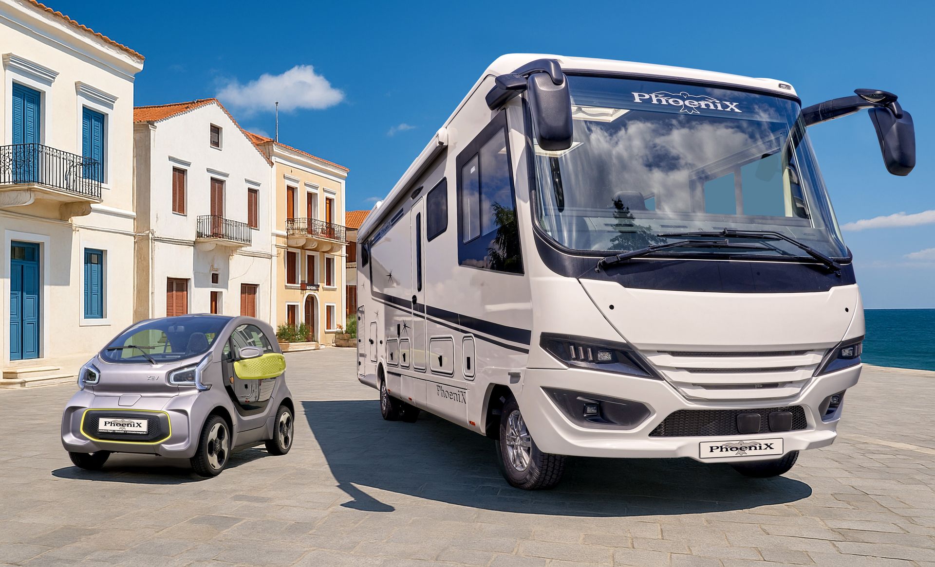 Wohnmobil und kleines Elektroauto parken auf einer gepflasterten Uferstraße vor Gebäuden und dem Meer unter blauem Himmel.