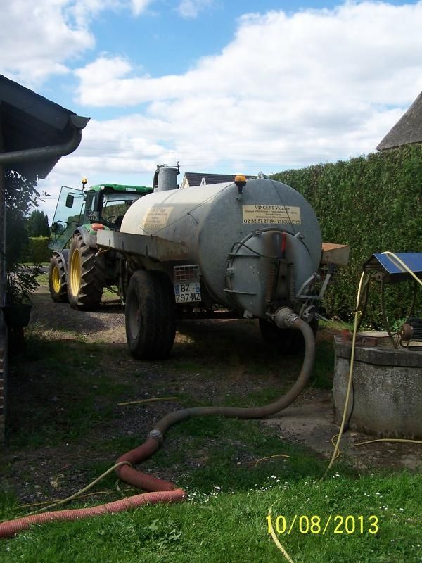 Tracteur avec remorque-citerne sur l'allée d'une ferme, tuyau de pompage au sol.