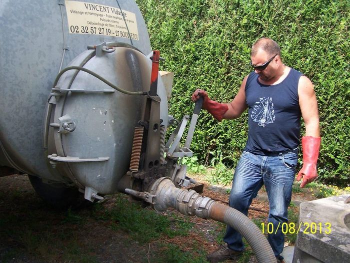Un homme manipule un gros tuyau relié à une cuve métallique à l'extérieur, avec une haie verte en arrière-plan.