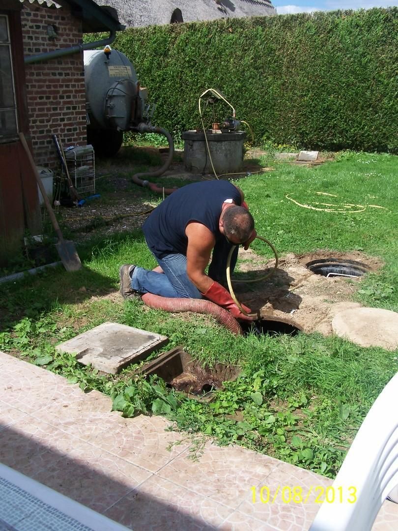 Une personne agenouillée près d'une bouche d'égout ouverte dans un jardin, travaillant avec des outils près de l'herbe.