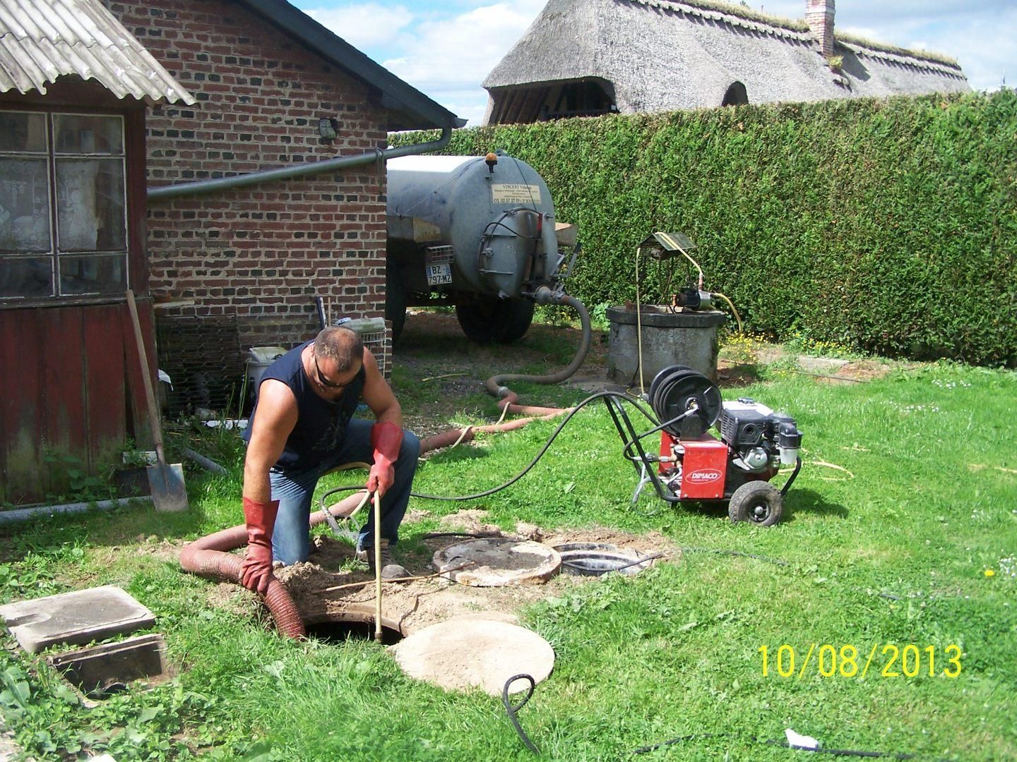 Une personne est accroupie au-dessus d'un trou dans un jardin, avec des outils et un générateur rouge à côté d'une maison en briques.