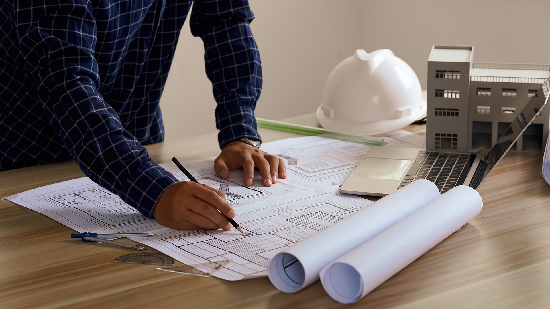 Arquitecto trabajando en planos en un escritorio con una computadora portátil, casco y maquetas de edificios.