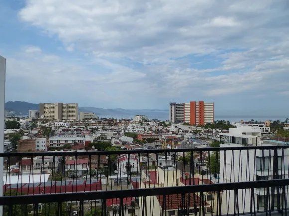 Cityscape with buildings under a cloudy sky, seen from a balcony with black railings.