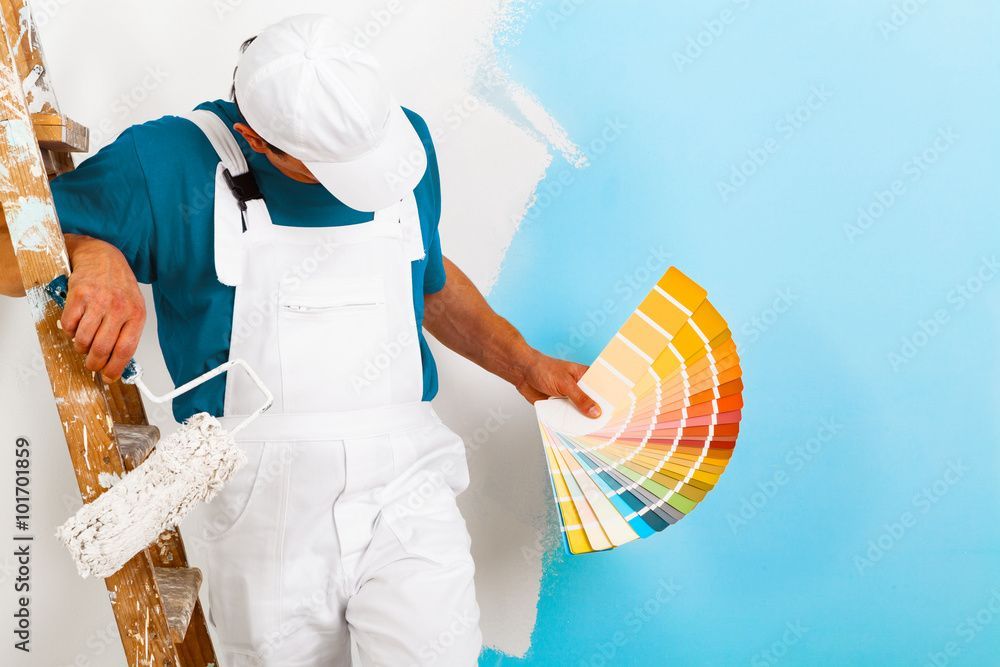 Maler in weissem Overall hält einen Farbfächer vor blauer Wand.