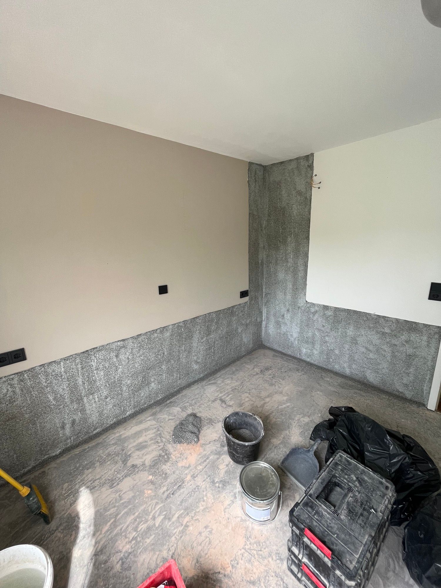 Ein Raum mit teilweise gestrichenen Wänden: Beige, Weiß und strukturiertes Grau. Bauwerkzeuge und Eimer auf dem Boden.