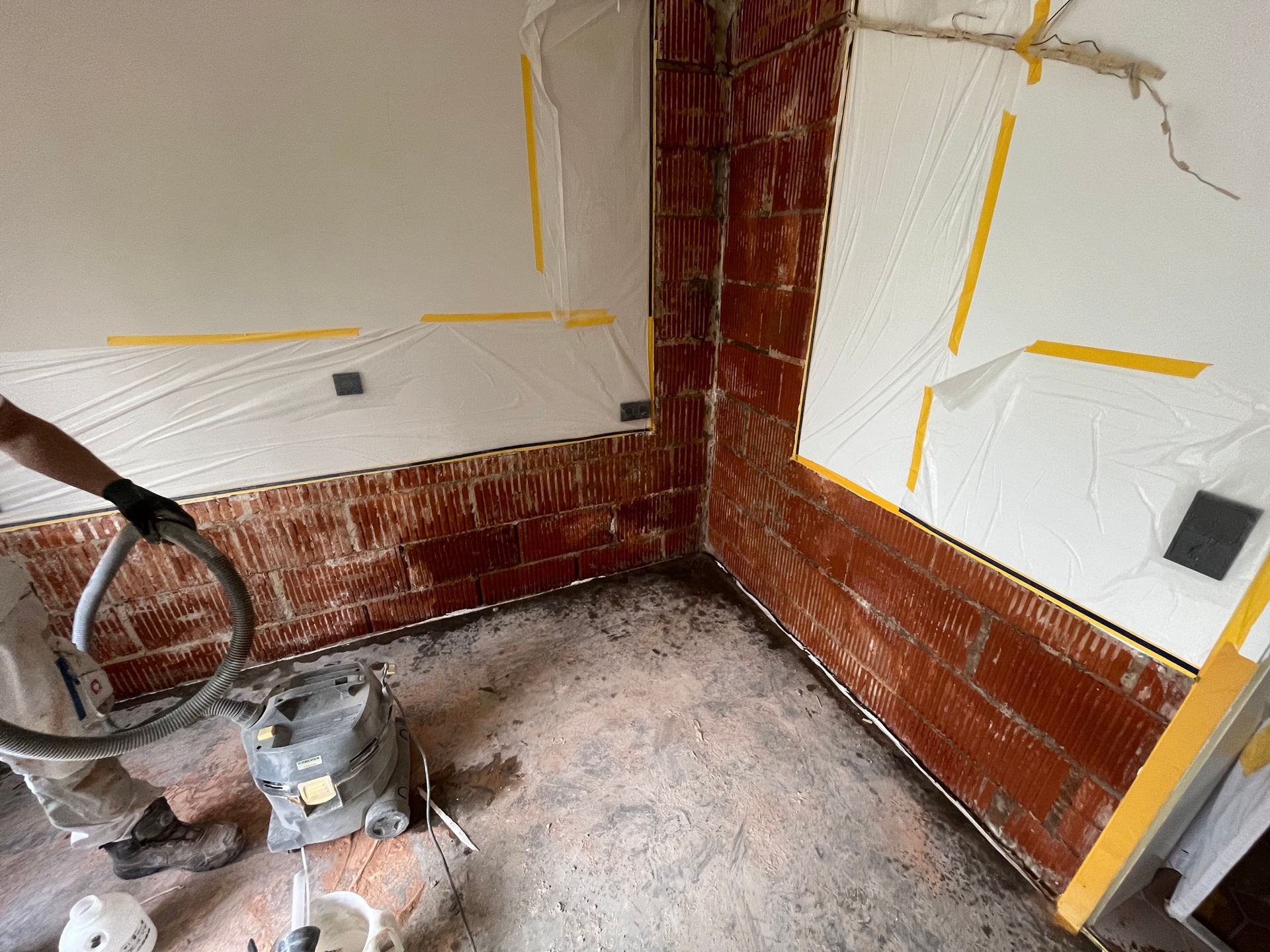 Ein Arbeiter saugt eine Innenecke einer Ziegelwand ab, Plastikfolie ist an den umgebenden Wänden festgeklebt.