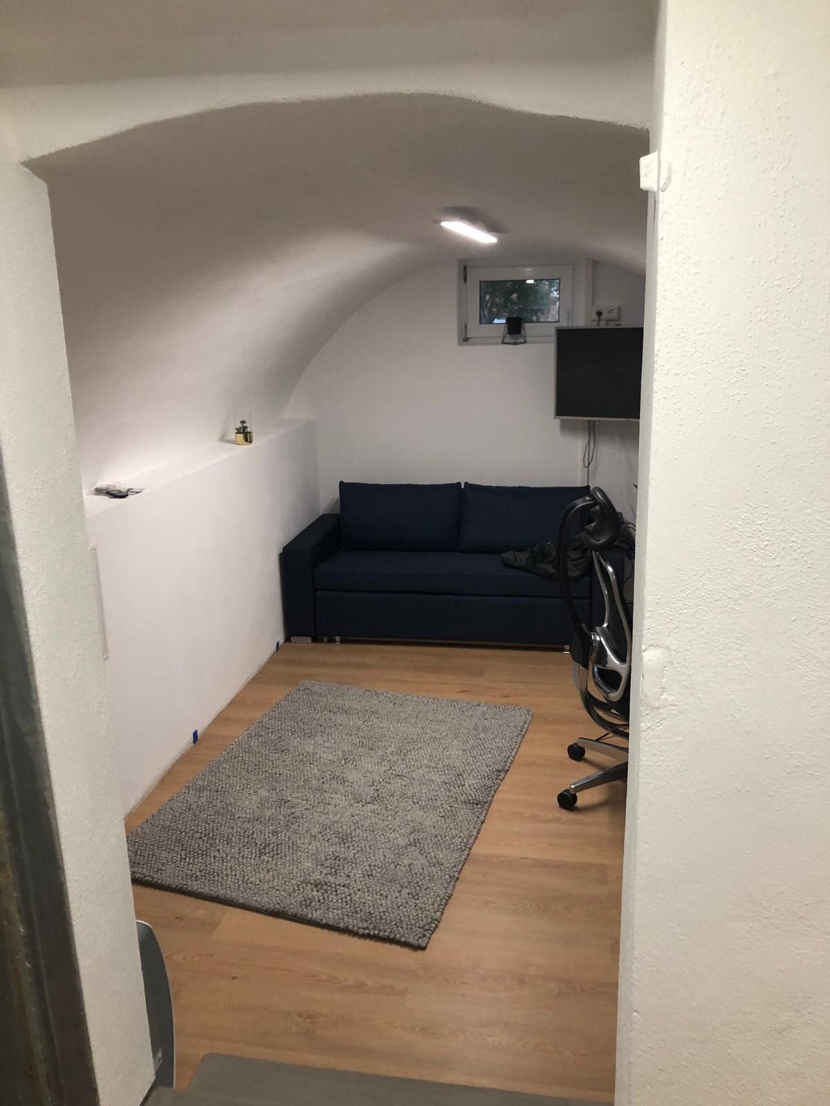 Kleiner, gewölbter Raum mit blauem Sofa, grauem Teppich, Fernseher und Schreibtischstuhl. Weiße Wände, Holzboden.