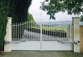 Portail d'allée en métal blanc à l'entrée d'une maison de banlieue, avec de la verdure et une route au loin.