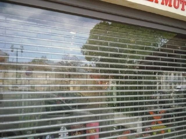 Façade fermée avec volet métallique, reflets d'arbres et de voitures garées.