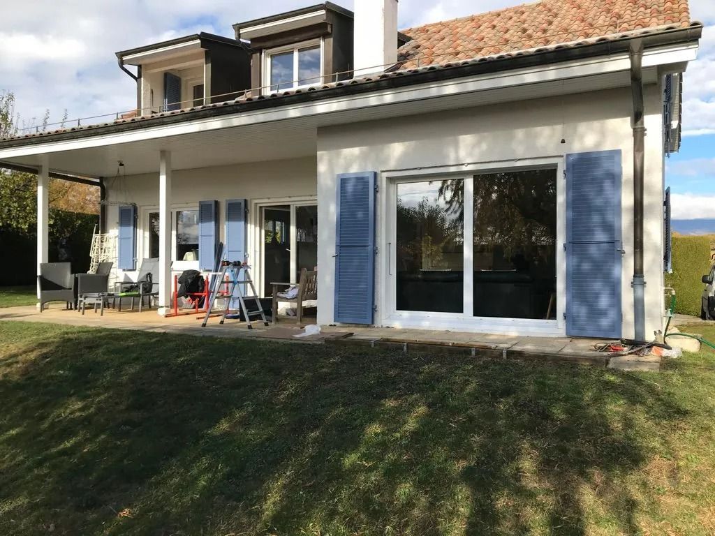 Terrasse arrière donnant sur la façade de la maison, grandes portes coulissantes en verre, volets bleus et pelouse devant.
