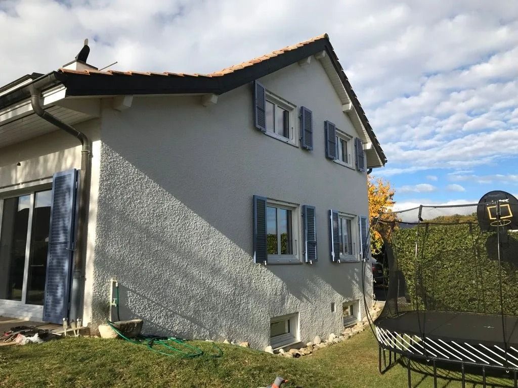 Maison grise à deux étages avec des volets bleus et un balcon dans une cour sous un ciel nuageux