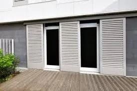Façade d'immeuble moderne avec trois portes en verre foncé et des volets gris clair au-dessus d'une terrasse en bois