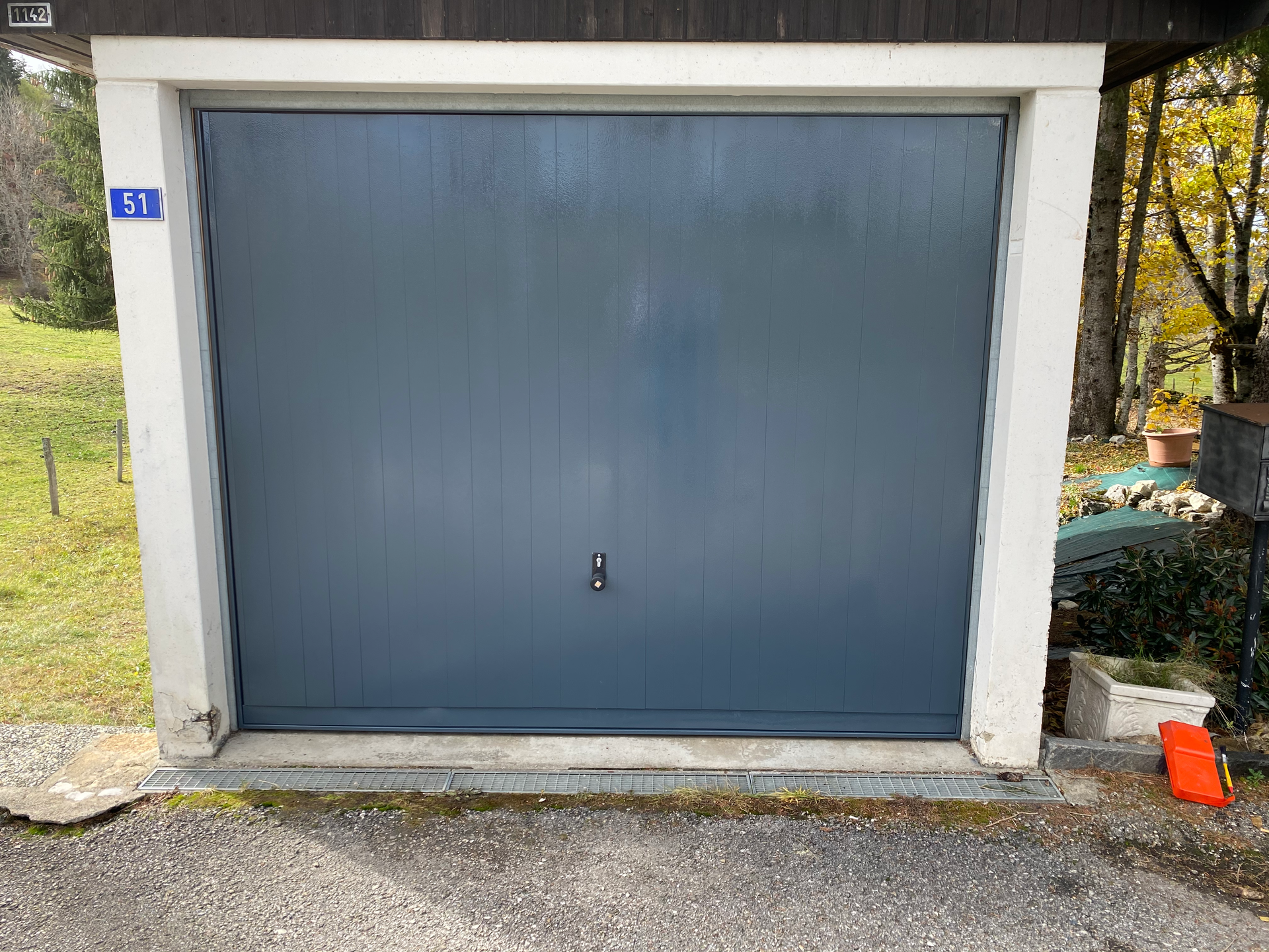 Porte de garage bleue avec encadrement blanc sur une allée en béton, avec un petit cône orange sur le bord droit.
