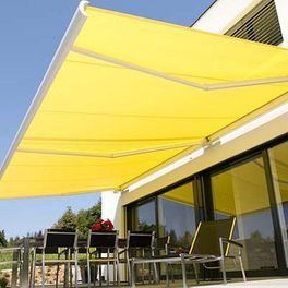 Un store rétractable jaune protège une terrasse extérieure avec des tables et des chaises près des portes vitrées.