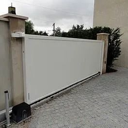 Portail coulissant blanc à côté de piliers beiges sur une allée résidentielle pavée.
