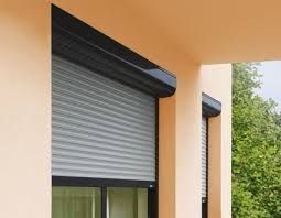 Volets roulants gris sur la façade beige d'un bâtiment, au-dessus des fenêtres, avec des arbres verts à côté.