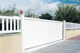 Portail coulissant blanc pour allée, adossé à un mur beige, avec une clôture blanche et des arbres en arrière-plan.