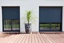 Deux fenêtres fermées aux volets sombres encadrent une grande plante en pot sur une terrasse en bois.