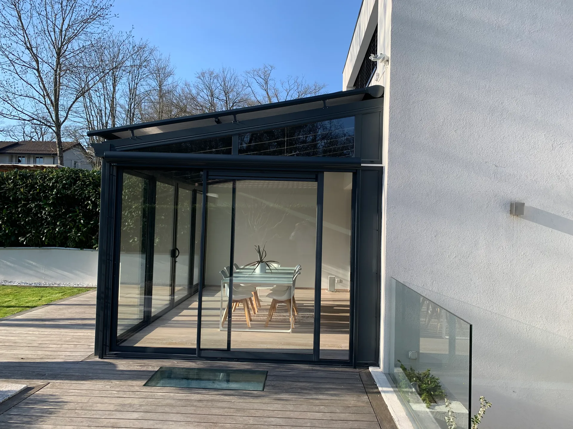 Extension vitrée donnant sur la terrasse d'une maison, avec une petite table et des chaises, baignée de lumière naturelle.
