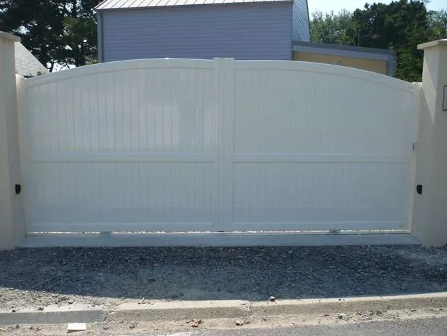 Portail d'allée en métal blanc fermé, à arc surbaissé, encastré dans un muret clair recouvert de gravier.