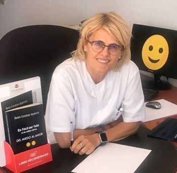 Una mujer está sentada en un escritorio con una cara sonriente en el monitor detrás de ella.
