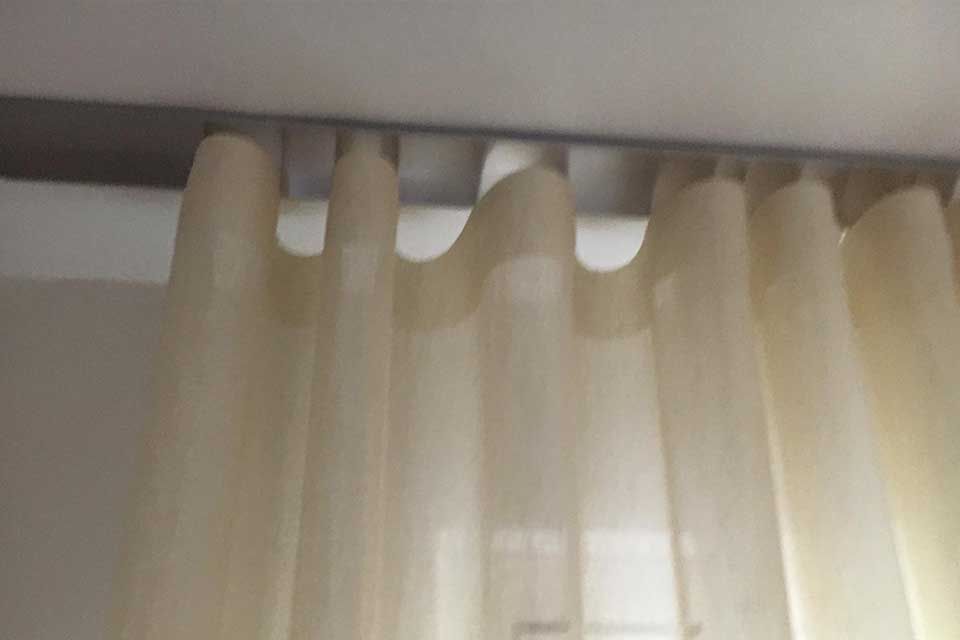 Beigefarbene, transparente Vorhänge hängen an einer grauen Schiene vor einer weißen Wand.