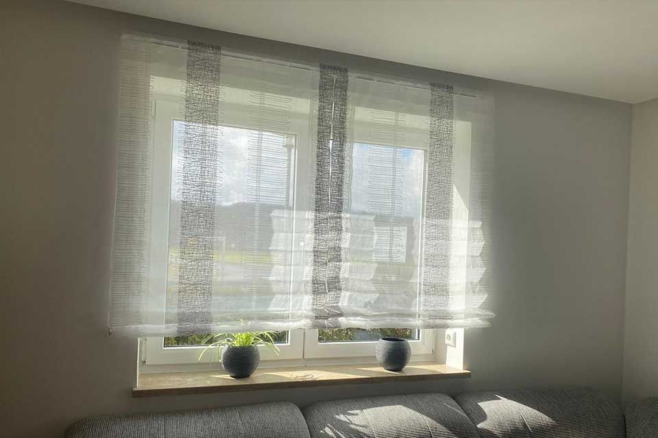 Fenster mit durchsichtigen weißen Jalousien, grau gemusterten Akzenten und durchströmendem Sonnenlicht.