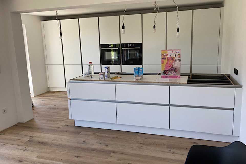 Moderne weiße Küche mit Kochinsel, Einbaugeräten und hellem Holzboden. Auf der Kochinsel liegen Bauwerkzeuge und eine Zeitschrift.