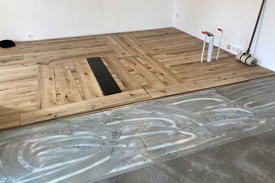 Holzbodenverlegung im Gange, mit Kleber auf dem Beton und verlegtem Parkett in einem Raum.