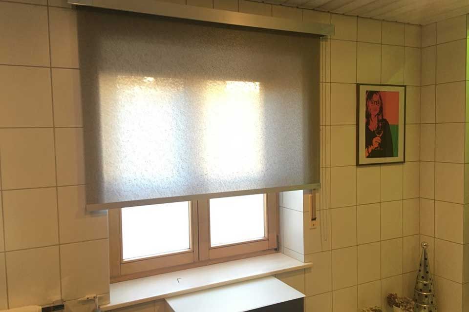 Fenster mit teilweise heruntergelassenem grauen Rollo, das Sonnenlicht hereinlässt. An der gefliesten Wand hängt ein gerahmtes Bild.