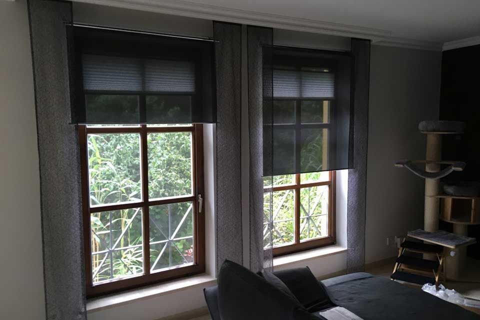 Zwei Fenster mit schwarzen Rollos und grauen Vorhängen; ein Bett und ein Katzenbaum im Zimmer.