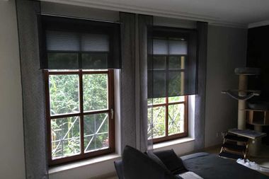 Zwei Fenster mit dunklen Jalousien und grauen Vorhängen. Ein Sofa ist zu sehen, in der Ecke steht ein Kratzbaum.