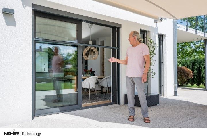 Mann öffnet automatische Glasschiebetür zur Terrasse. Er spiegelt sich im Glas. Modernes weißes Haus außen.