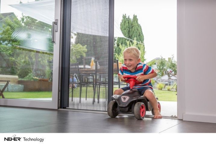 Ein kleiner Junge fährt in einem Spielzeugauto im Haus auf eine Glastür mit einem Fliegengitter zu, im Hintergrund ist draußen etwas zu sehen.